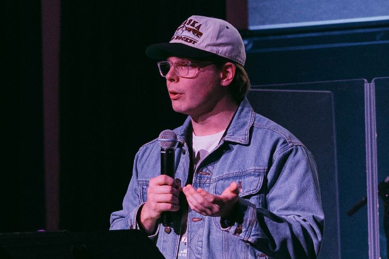 Man in glasses, denim jacket, and hat speaks into a microphone on a stage.