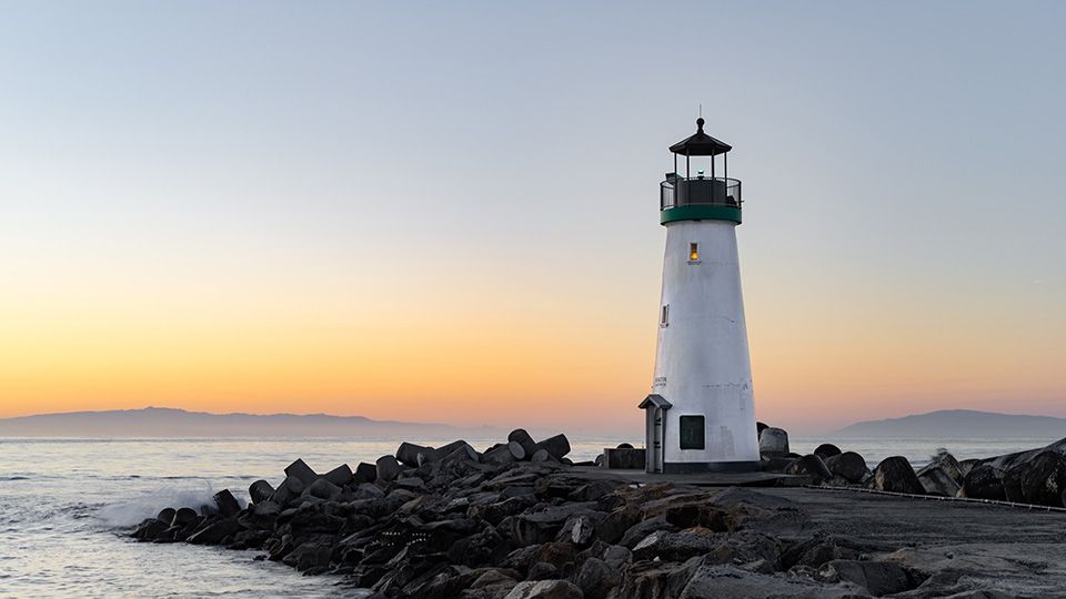 White lighthouse on rocky shore at sunset, orange and blue sky.