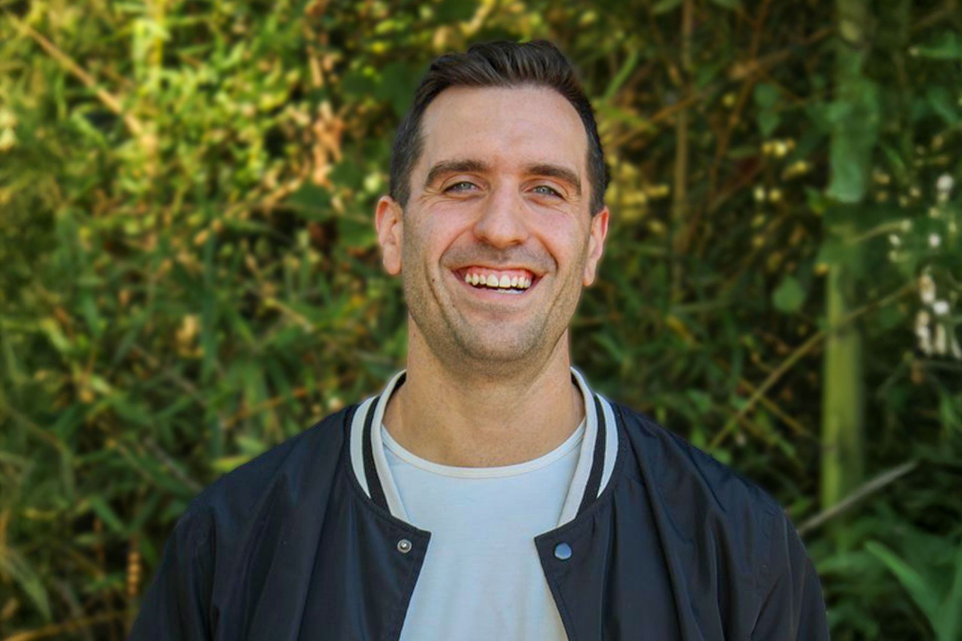 Man smiling broadly, wearing a black bomber jacket over a white shirt, against a green leafy backdrop.