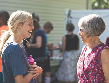 Two women conversing outdoors, smiling. Others in the background. One holds a water bottle.