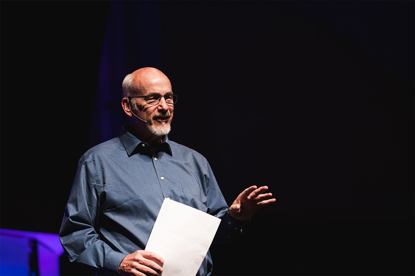 Man in blue shirt, eyeglasses, gesturing, holding paper, speaking on stage.