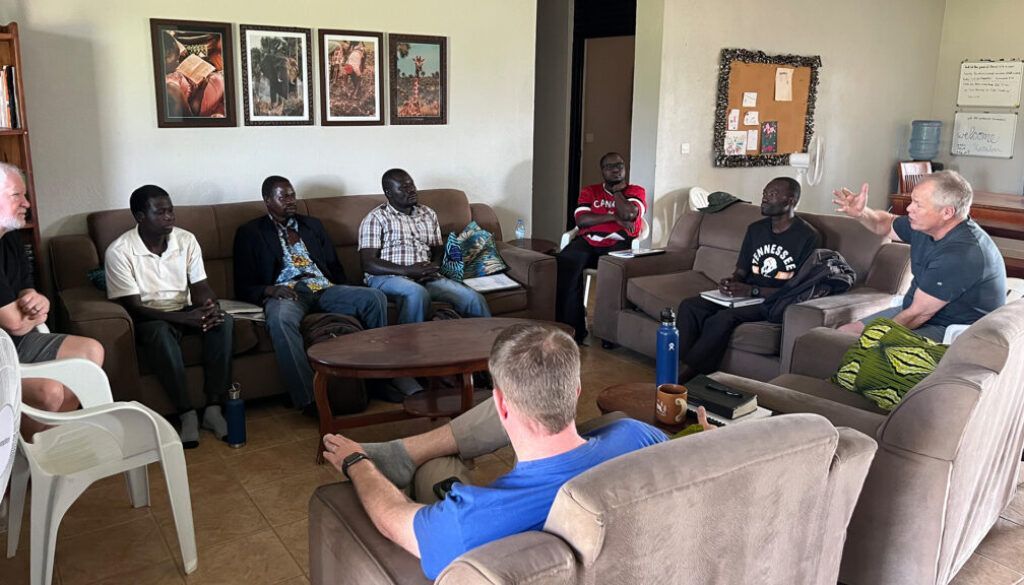 People sitting in a living room, some on sofas, engaged in a group discussion.