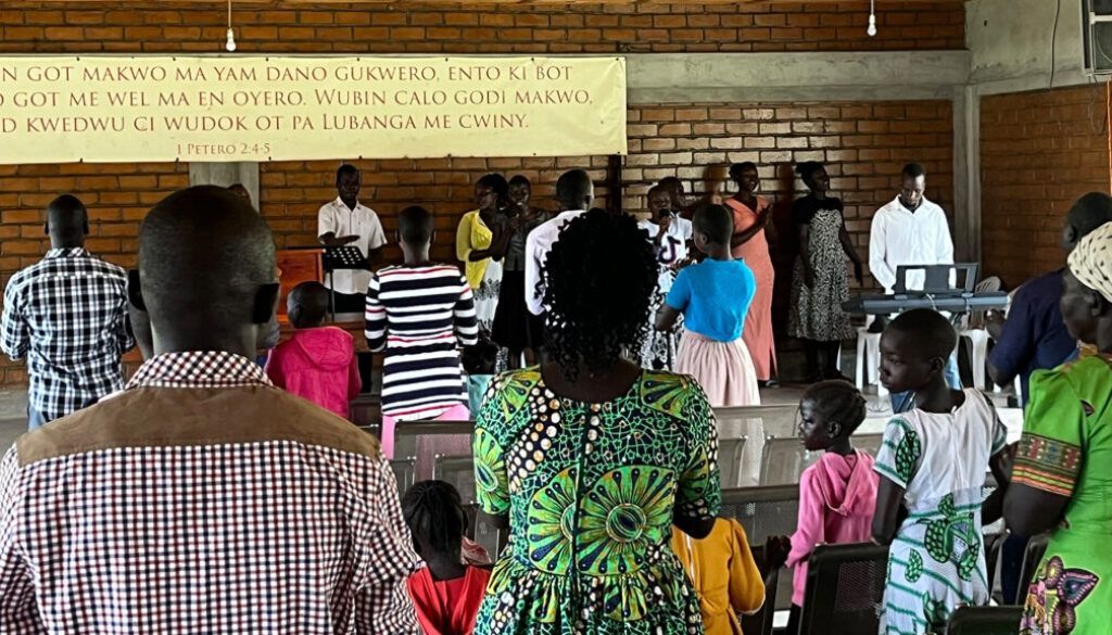 People singing in a room, with a banner above. Some are standing, others are looking down.