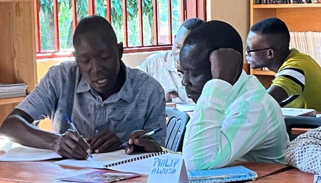 Men writing in notebooks at a table, sunny room, focused expressions.
