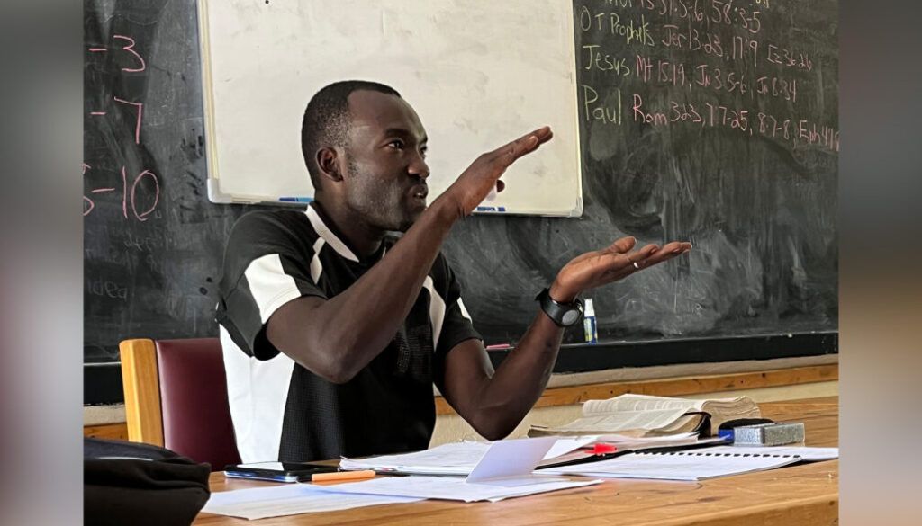 Man gesturing in front of a chalkboard. He is wearing a black and white shirt and sitting at a desk with papers.