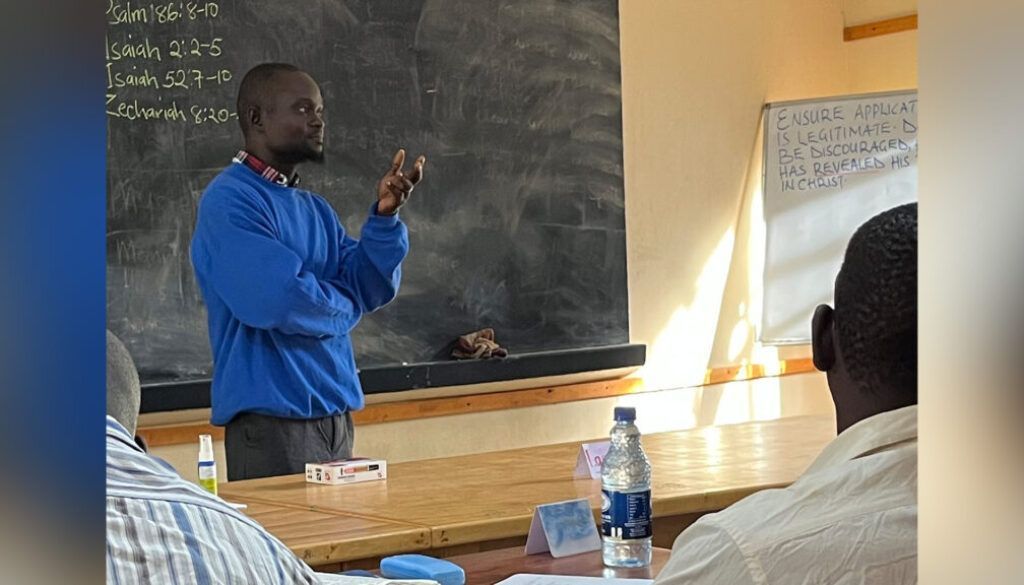 Teacher in blue sweater gestures in classroom, blackboard behind.