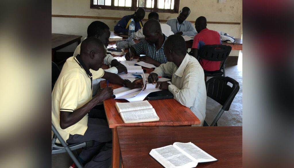 post-6 copy Group of men seated at tables, reading and writing in a room with a window.