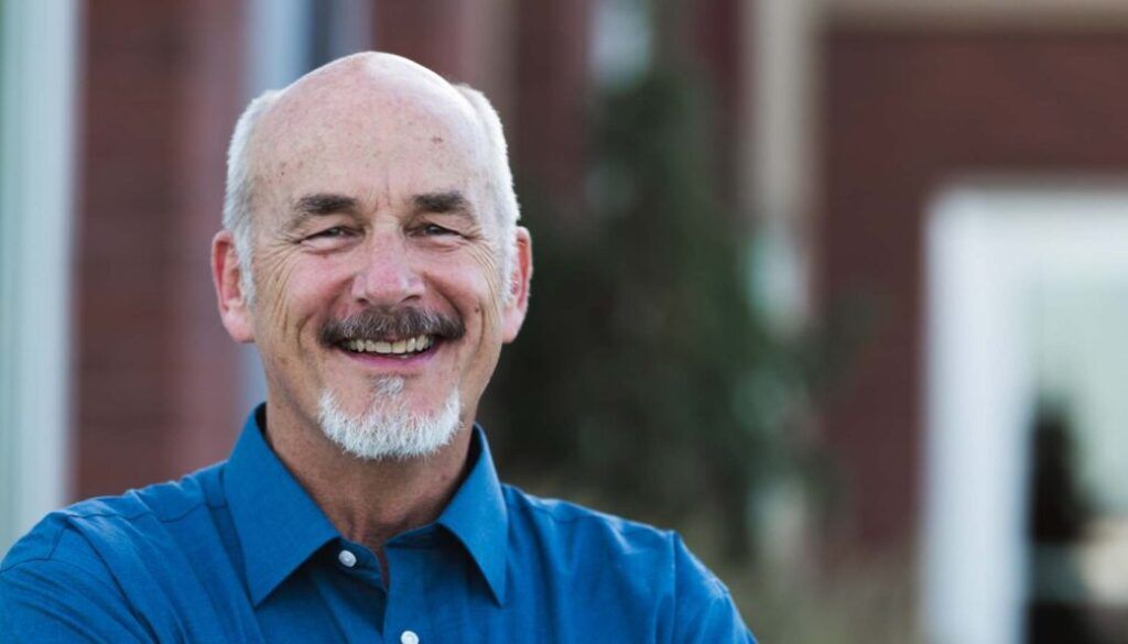 Smiling man with white beard and blue shirt, outdoors near brick building.