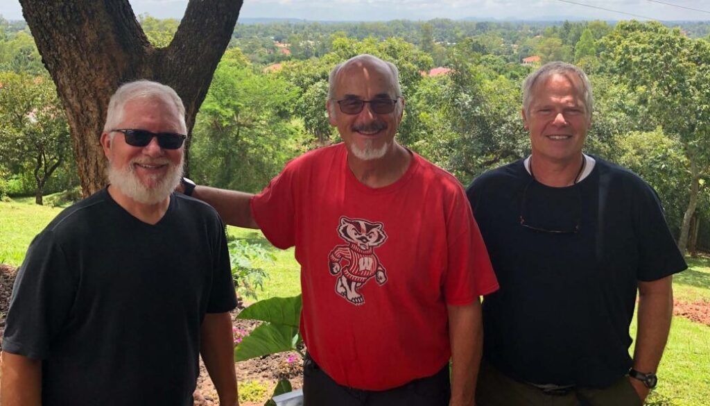Three men smiling outdoors, arms around each other, in front of a green landscape.