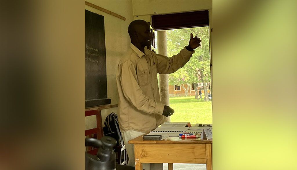 Man gesturing with his right hand in a classroom setting, smiling, standing near a table and open window.