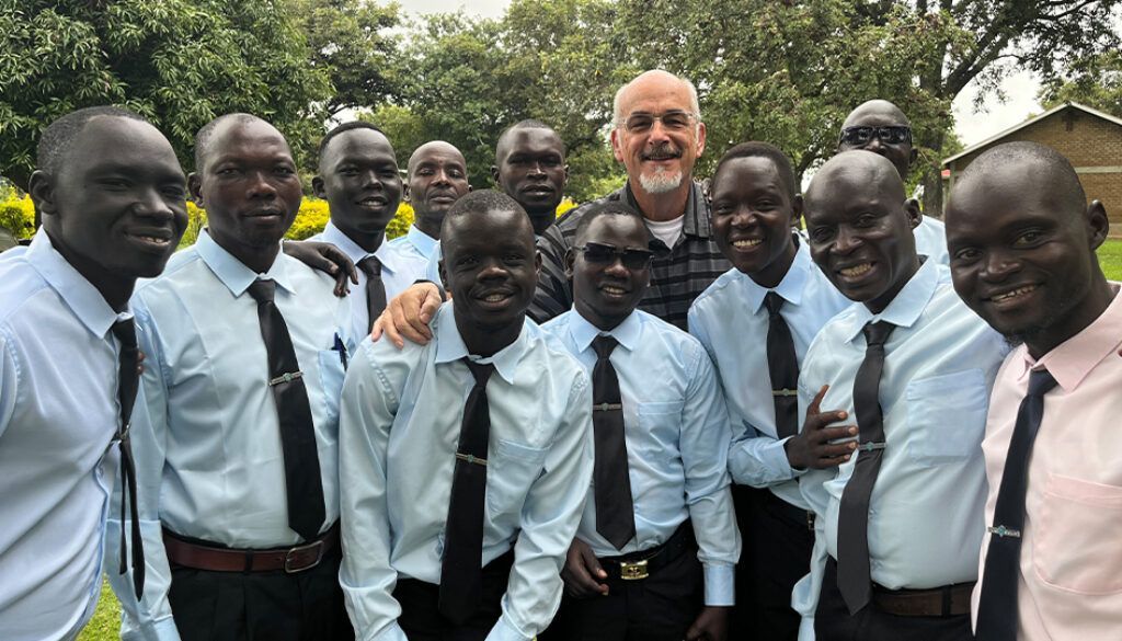 Group of men in light blue shirts and black ties smiling outdoors with a man with a beard.
