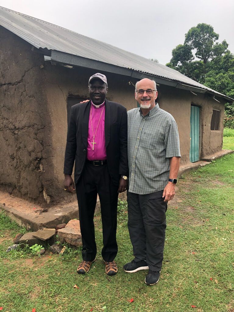 Two men stand in front of a small, rustic building. One wears a clerical collar, the other a button-down shirt.