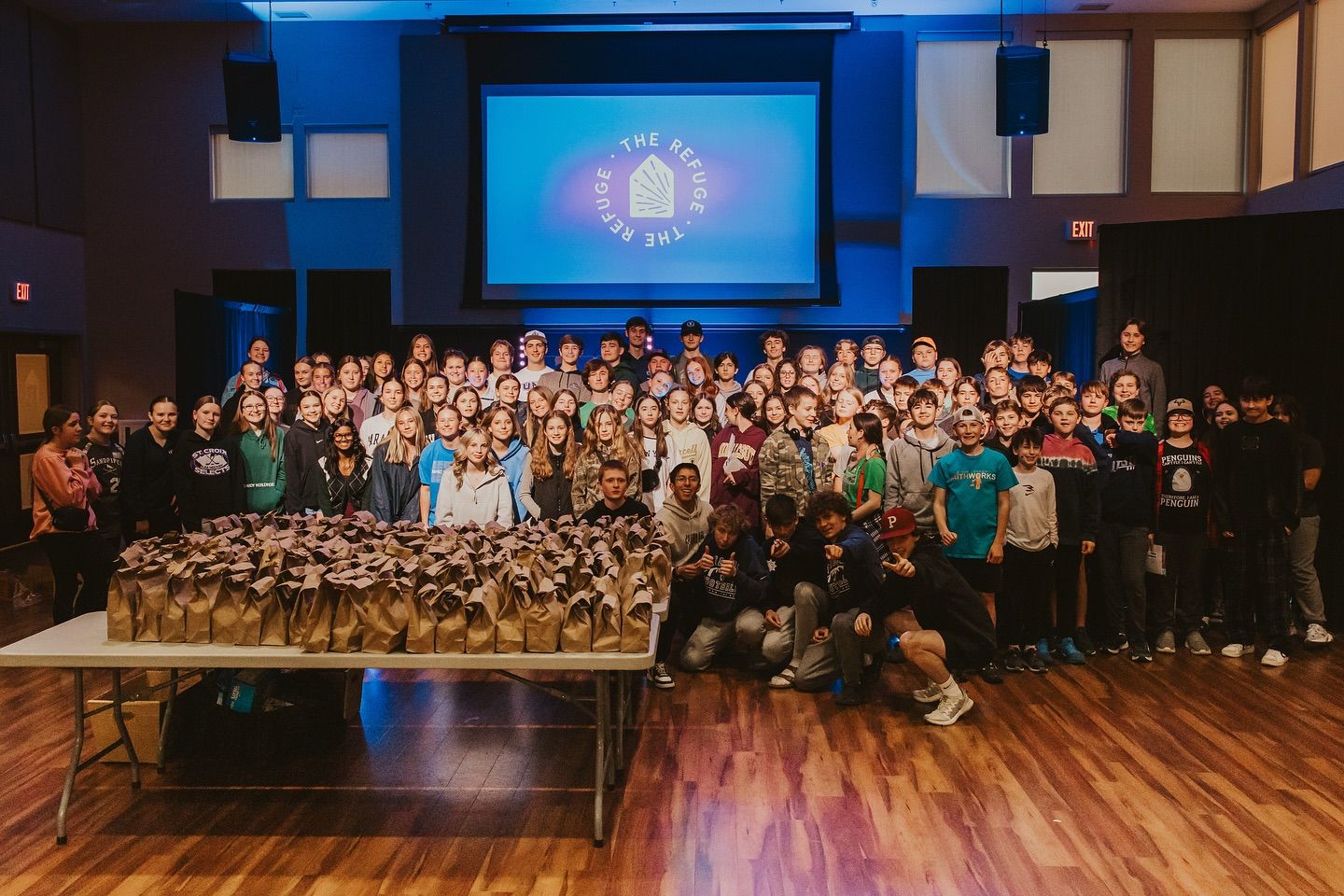 Large group of people pose with bags in front of a screen with a logo in a wooden-floored room.
