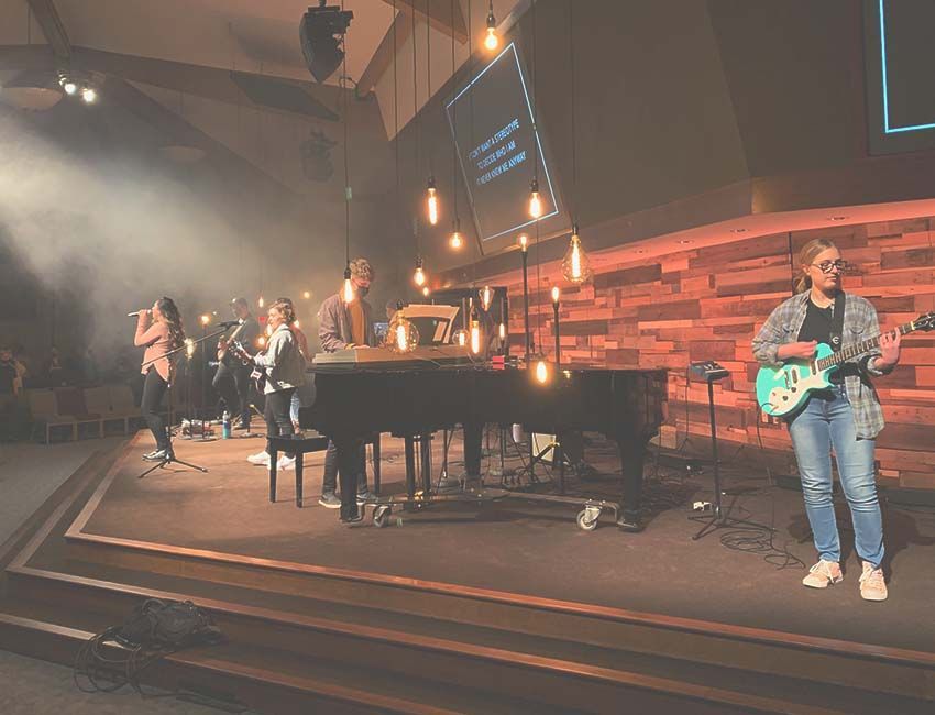 A band performs on a stage with a piano, guitar, and vocalists under warm lights.