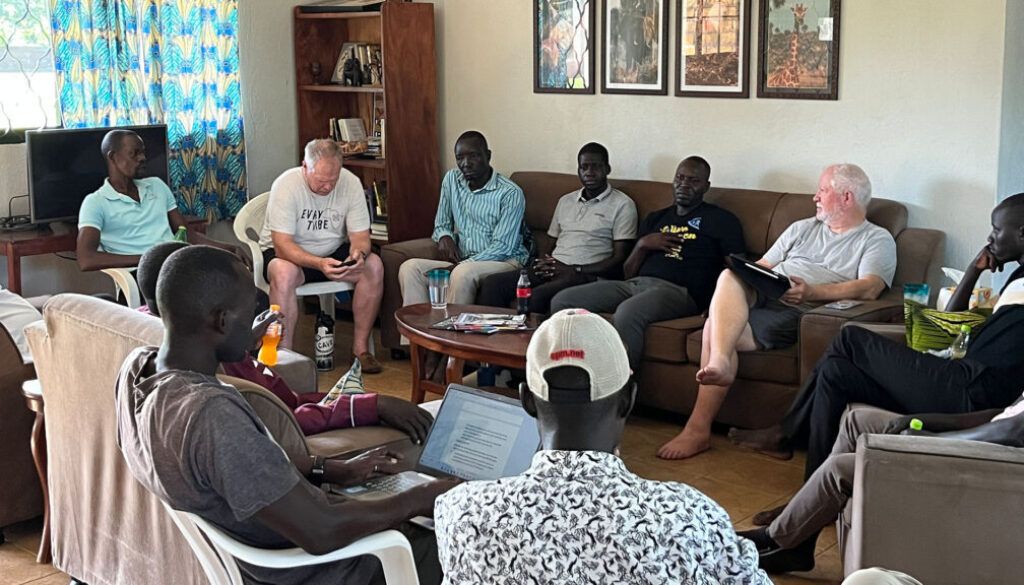 Group of people seated, some on a sofa, in a room. Some are looking at a laptop.