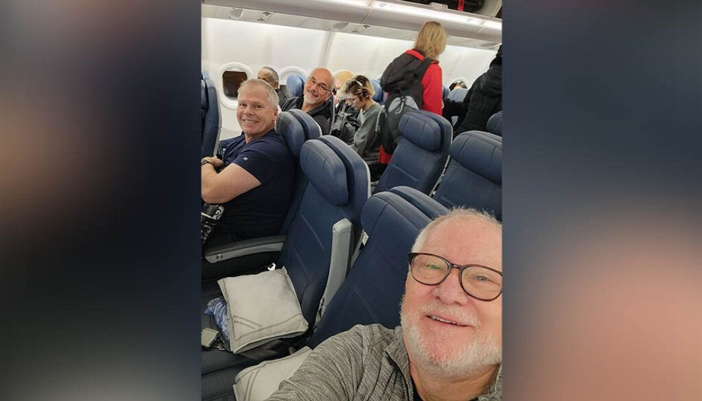 uganda11post2featured Two men smiling for a selfie on an airplane; passengers in background.