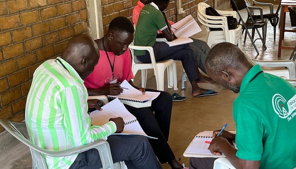 Several people sitting and writing on papers in a room with a brick wall.