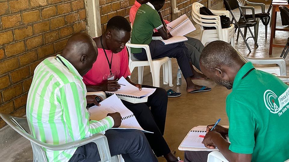 Group of people writing on paper in a room; one wearing green shirt, pink shirt, others in the background.