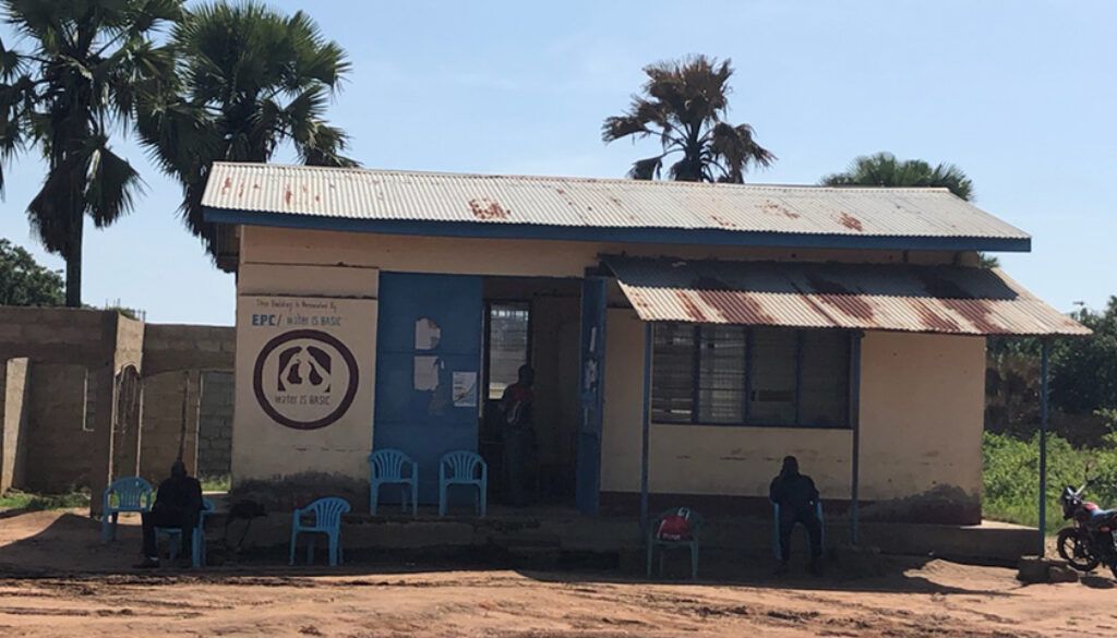 Small, white building with blue door and chairs outside. Rusted awning, sign with logo, and two people seated.