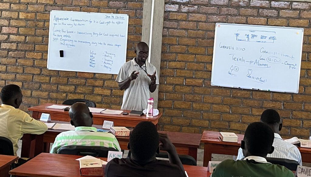 ugandapost3featured A teacher lecturing in front of a class. Whiteboards behind him, students seated at desks.