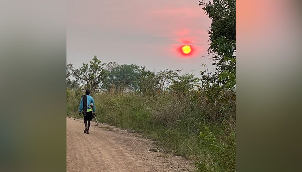 ugandapostimg Person walking down a dirt road towards a vibrant orange sun setting over grassy field.