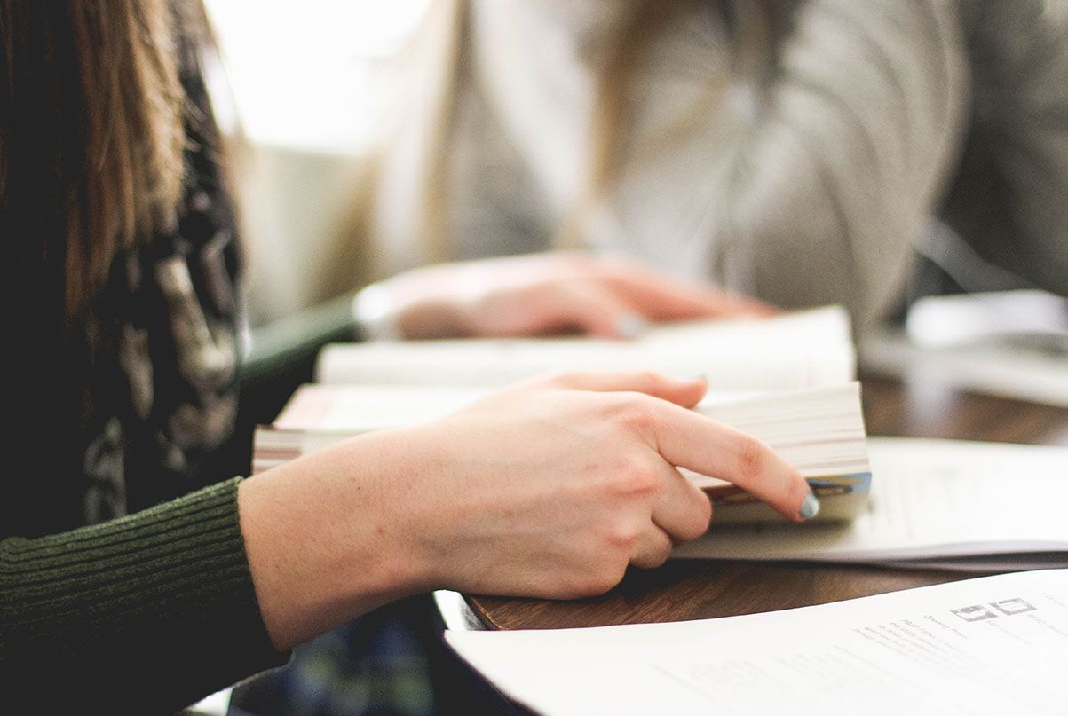 Person's hands turning pages of a book on a wooden table. Another person's hands are out of focus in the background.