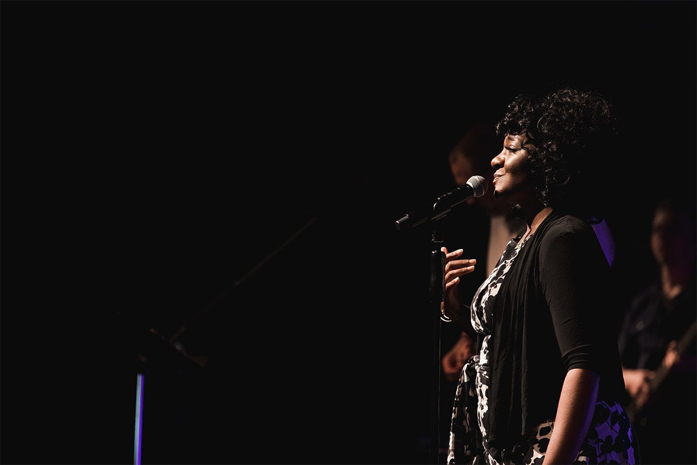 Woman singing into a microphone on stage, wearing a black and white patterned dress, with musicians in the background.