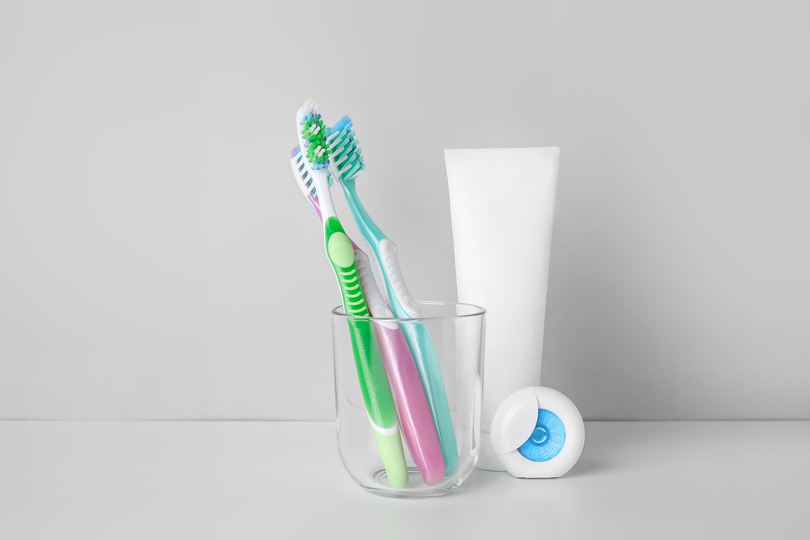 Two toothbrushes in a glass, toothpaste tube, and floss on a white surface against a gray background.