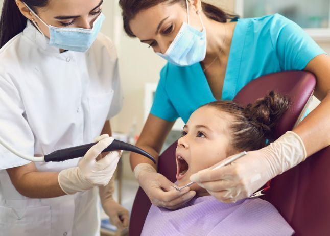 Dentists examining a young girl's teeth. Two adults in masks and gloves. Girl in dentist chair, mouth open.