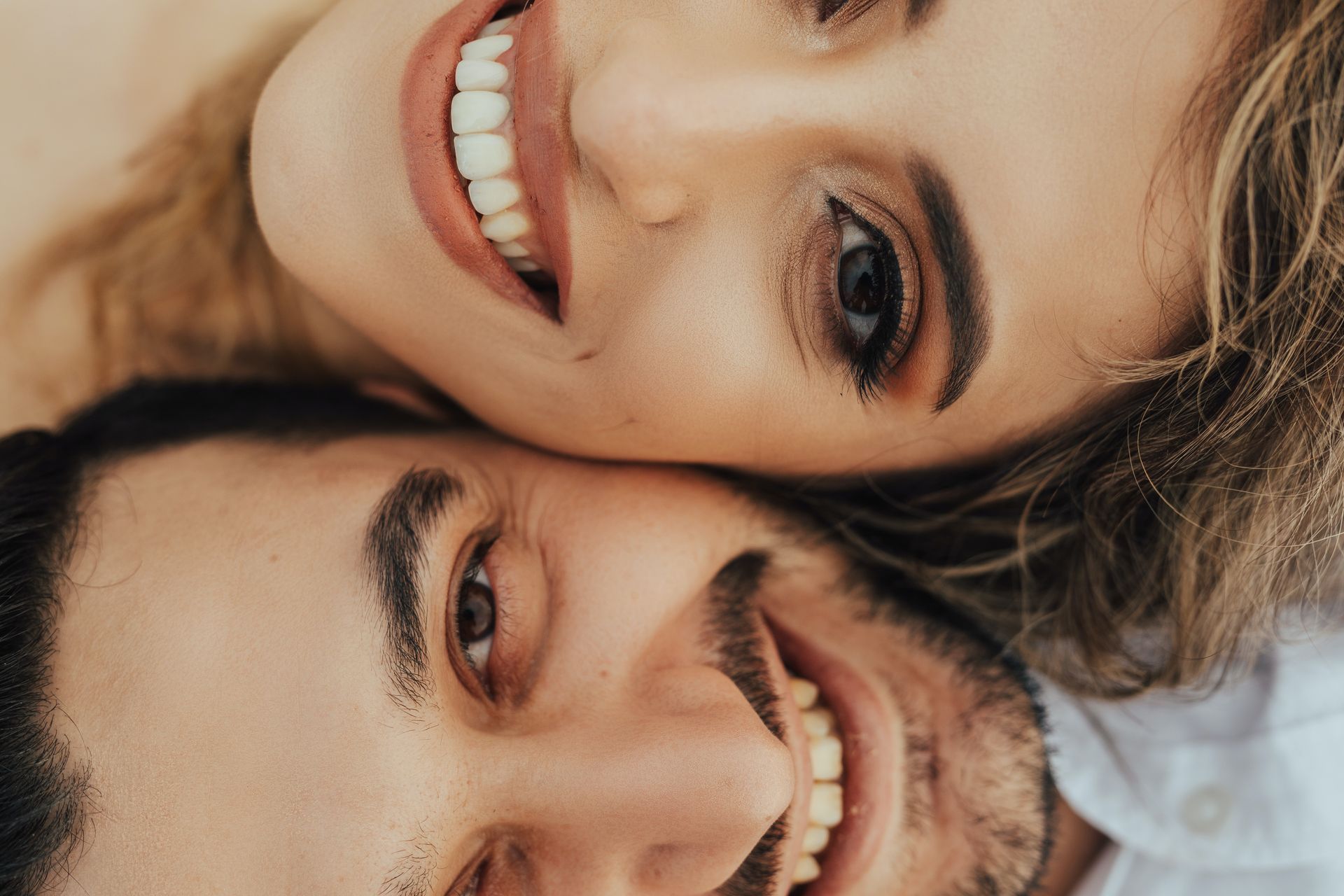 Two smiling faces close together, one with curly hair and one with a mustache, against a light background