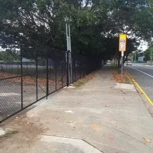 A sidewalk next to a black chain-link fence, a 40 km/h speed limit sign, and a road with trees in the background.