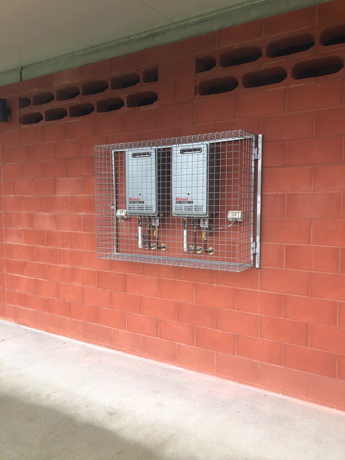 Two tankless water heaters mounted on a red brick wall, enclosed within a protective metal security cage.