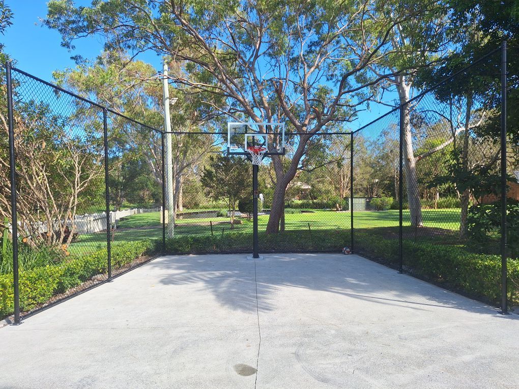 An outdoor basketball court with a hoop and black mesh fencing surrounded by green trees and bushes on a sunny day.