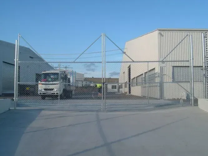 A white truck exits through a large chain-link metal gate onto a concrete lot between two industrial buildings.