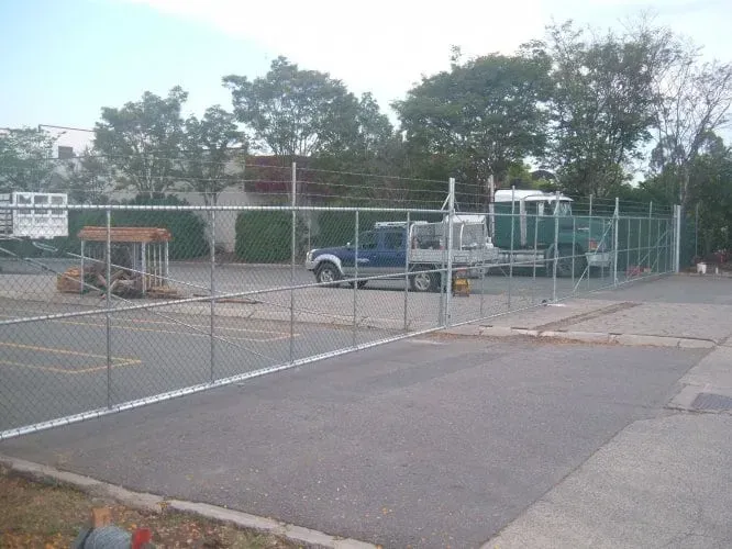 A fenced-in parking area featuring a blue pickup truck and a larger truck in an outdoor lot with trees in the background.