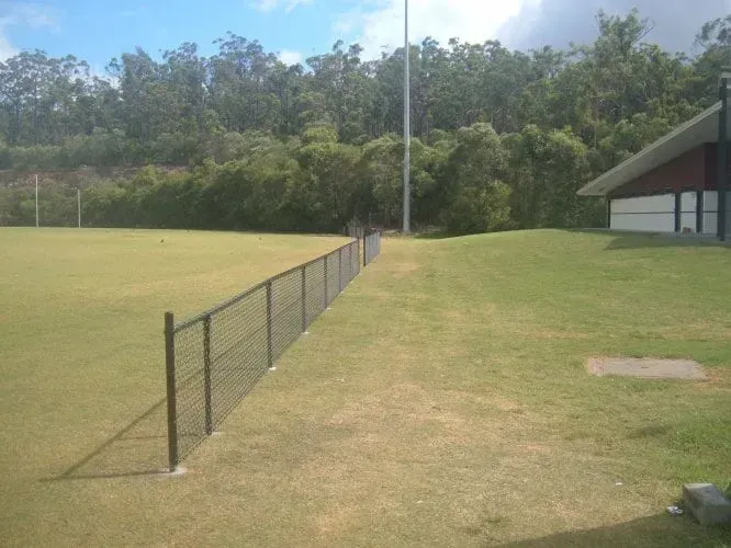 A chain-link fence divides a grassy sports field from a grassy lawn area near a building under a bright blue sky.