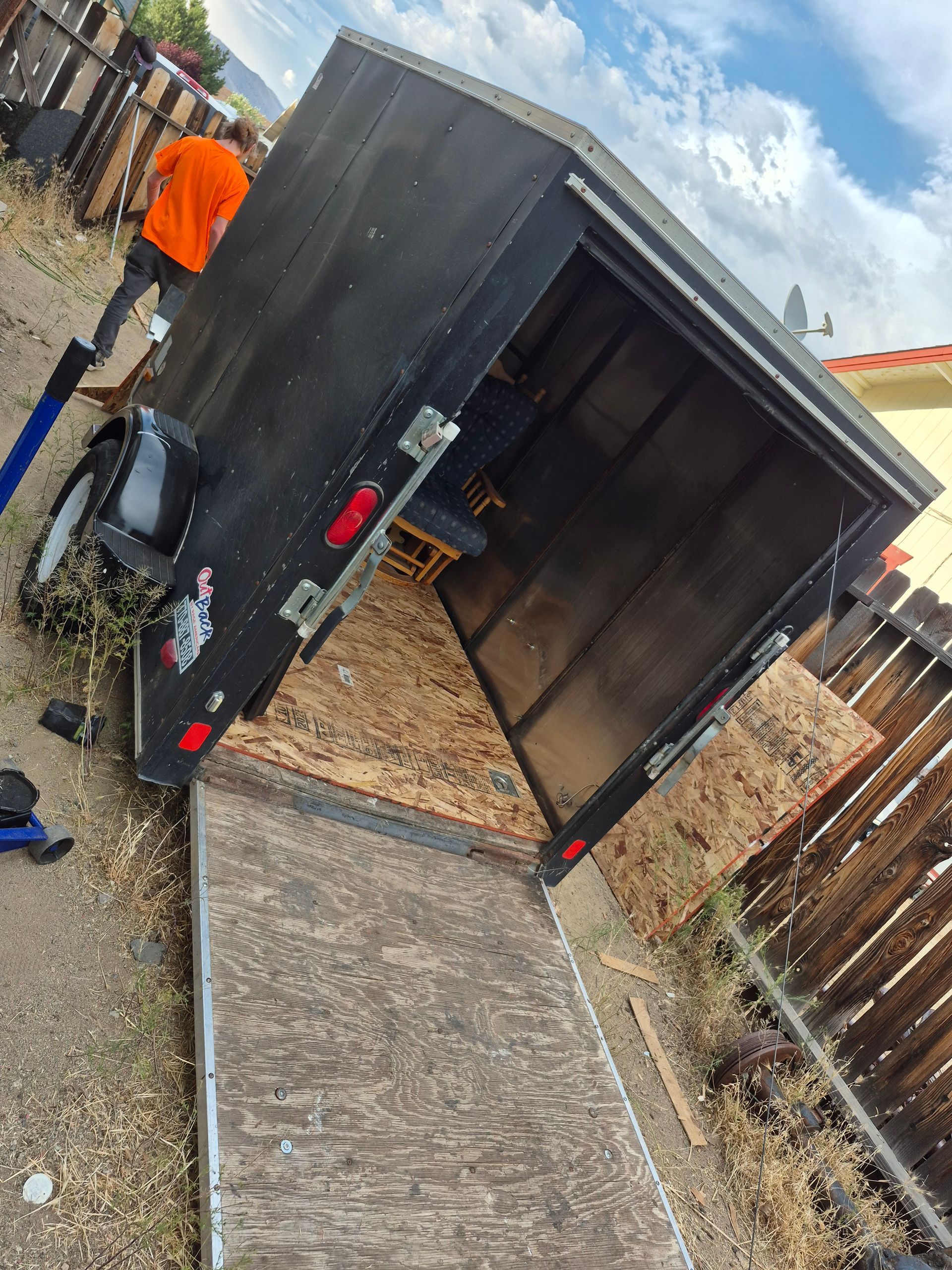 Black trailer with ramp down, person in orange shirt standing near it, outdoors.