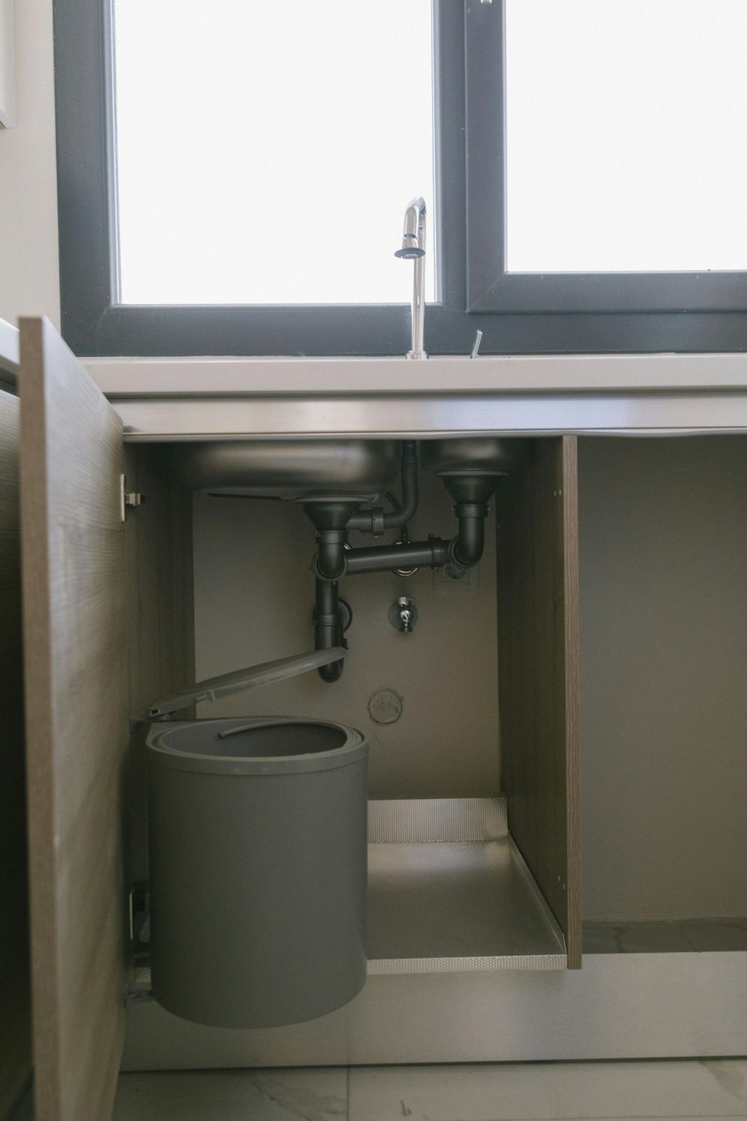 Kitchen sink cabinet with plumbing, a pull-out trash can, and a window in the background.
