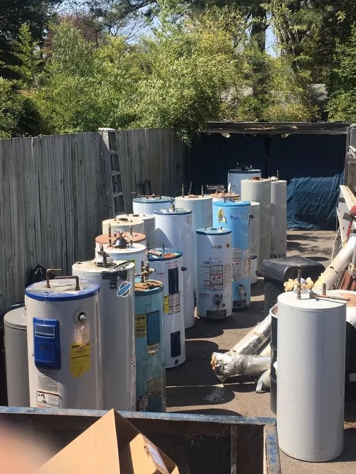 Row of white water heaters and pipes stored in a fenced outdoor yard