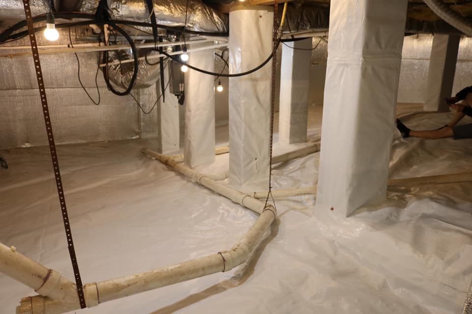 Crawl space with white plastic sheeting, support columns, pipes, and a hanging lightbulb.