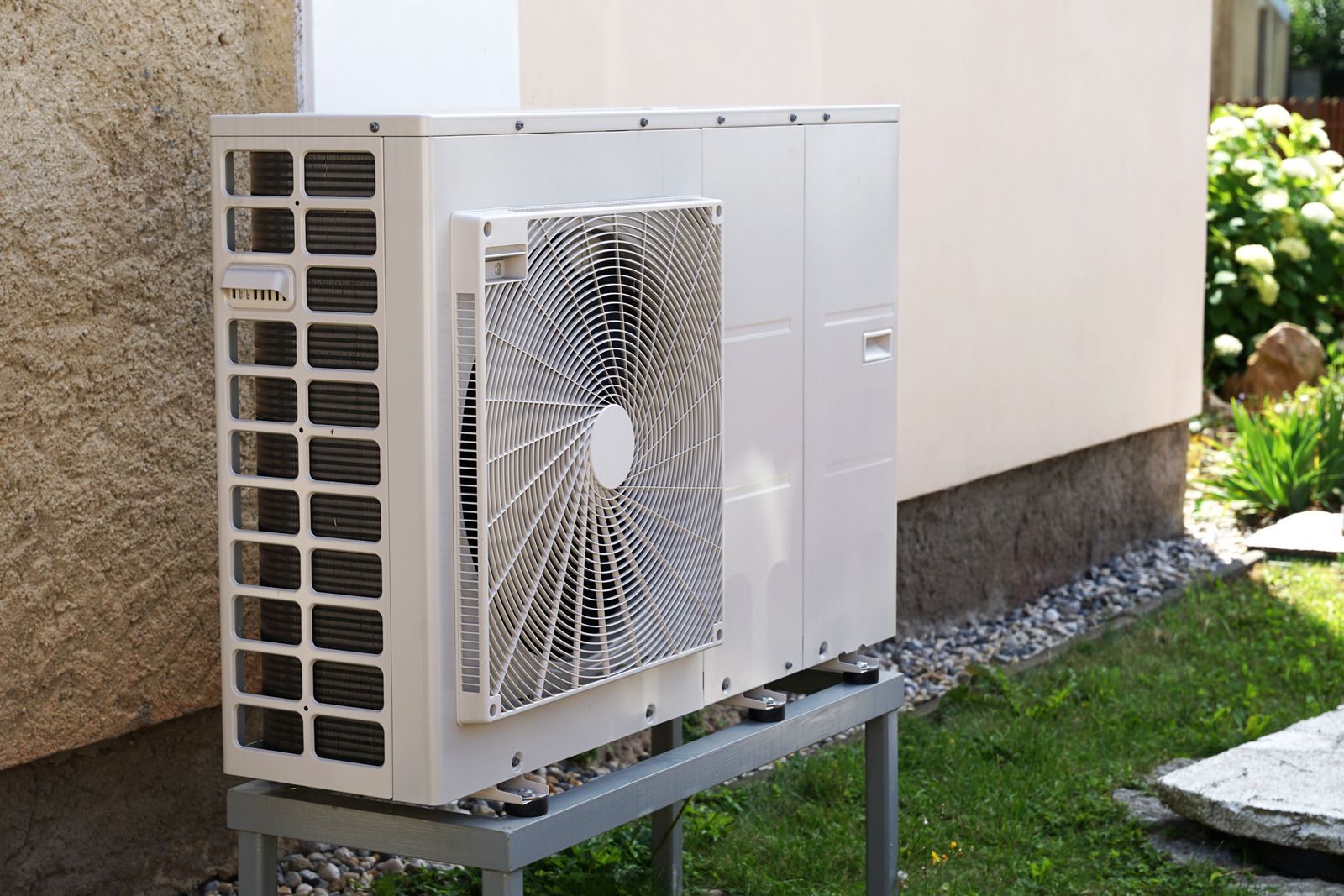 Outdoor air conditioner unit beside a building wall on a metal stand, with a fan grille and sunlight