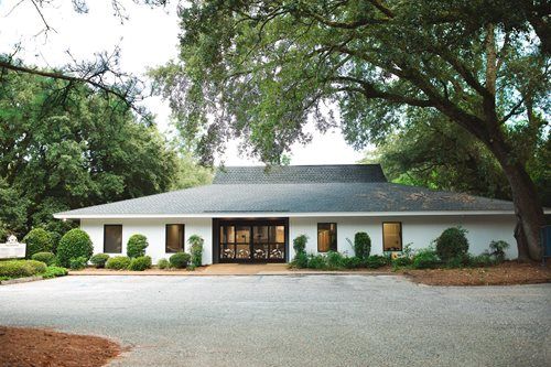 White single-story house with dark roof and large trees in front, seen from a gravel driveway