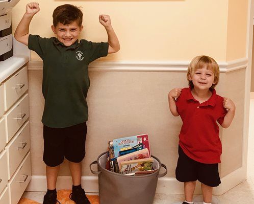 Two children standing by a hallway wall, smiling and flexing beside a bucket of books and toys