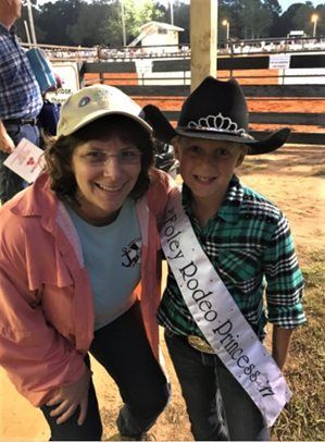 Two smiling people in cowboy hats at an outdoor rodeo, one wearing a “Little Dude” sash.