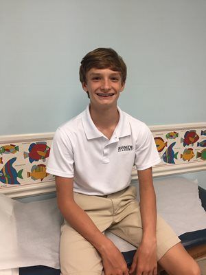 Smiling boy sitting on a bed in a white polo and khaki shorts, with colorful wall border behind him.