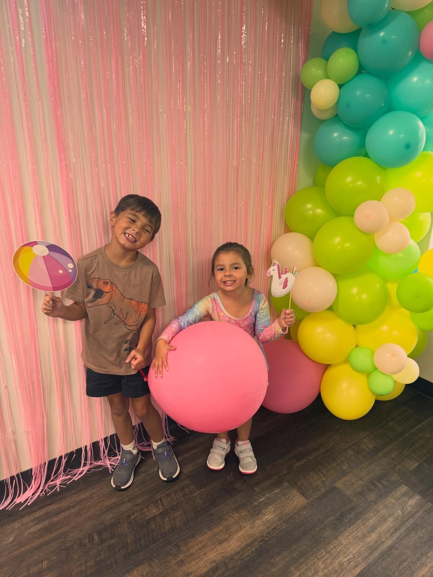 Two children standing by a hallway wall, smiling and flexing beside a bucket of books and toys