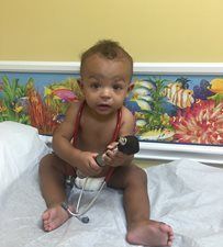 Young child sitting on a hospital bed holding a blood pressure cuff, with a colorful mural behind.