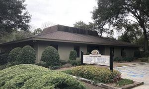 Low building with manicured shrubs and a sign at the entrance, surrounded by trees on an overcast day