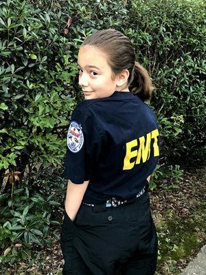 Child in a navy jacket with yellow letters glancing back while standing beside green bushes.