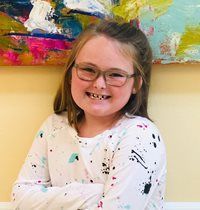 Smiling person with glasses and freckles in a white shirt, standing before colorful abstract artwork